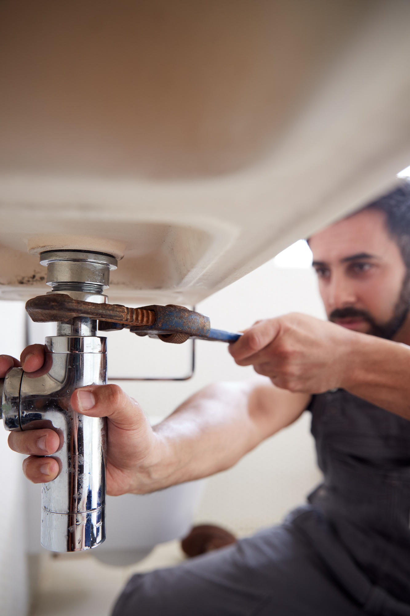 Male Plumber Using Wrench To Fix Leaking Sink In Home Bathroom